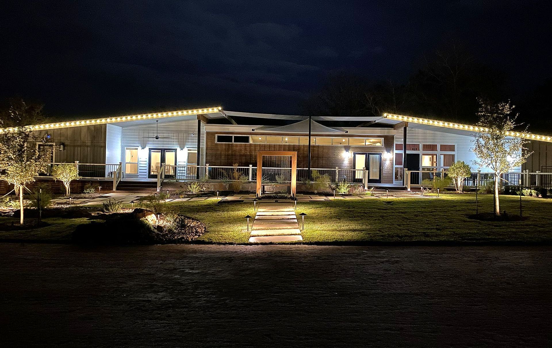 Night view of a modern home with lit trees and walkway, string lights on the roof, and interior lights visible.