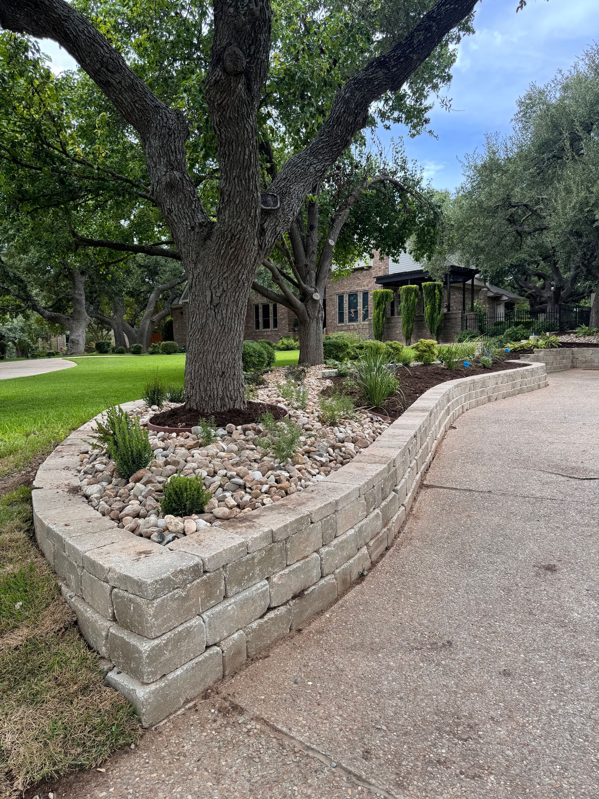 Stone retaining wall borders a flower bed around a tree, next to a driveway; house in background.