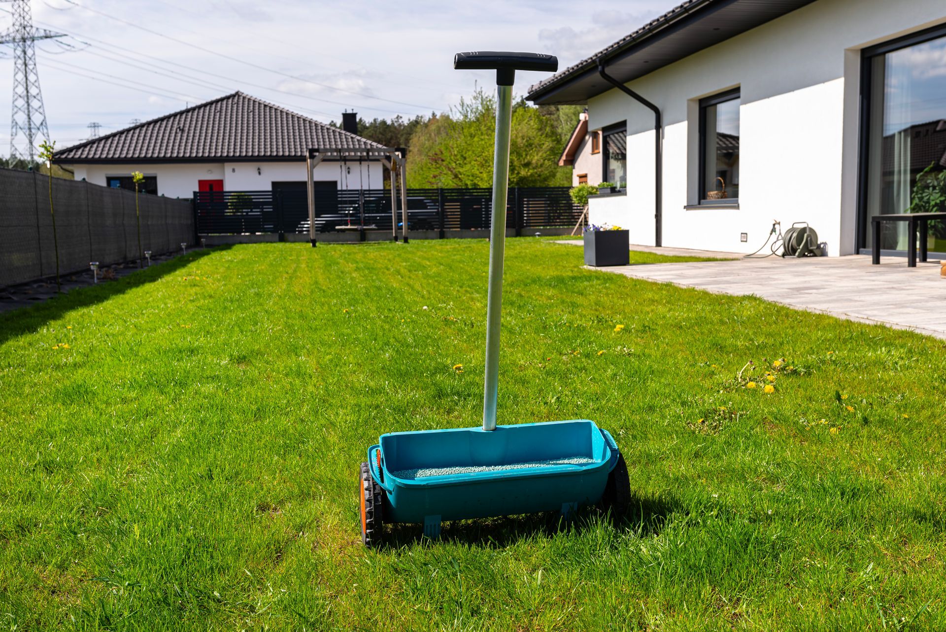 Lawn spreader in a green yard, with a house and fence in the background.