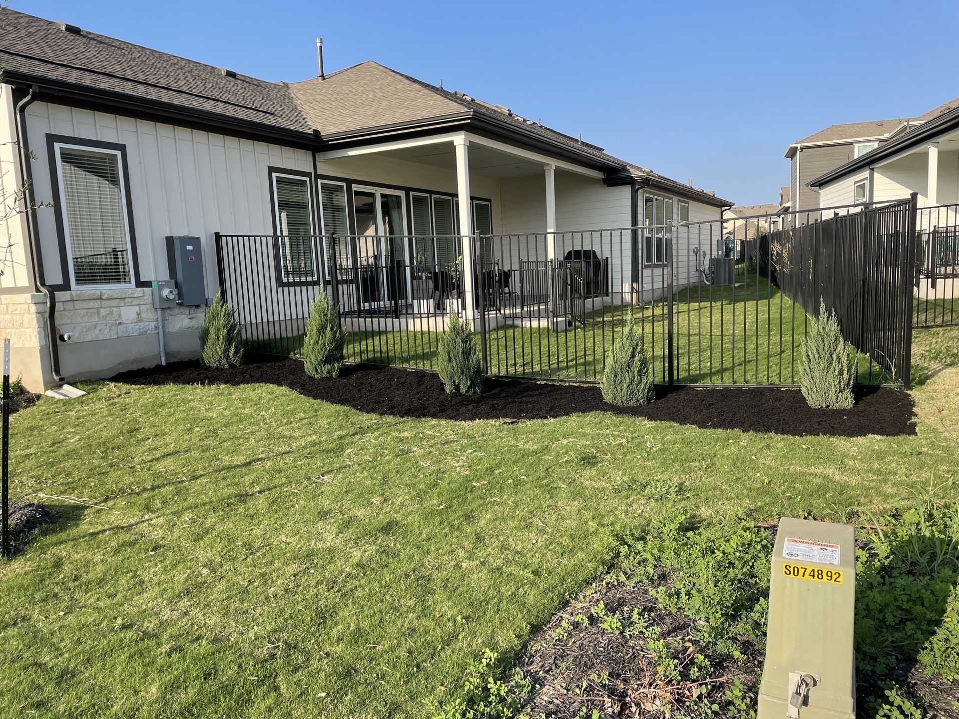 Backyard with a white house, patio, black fence, trees, and green grass.