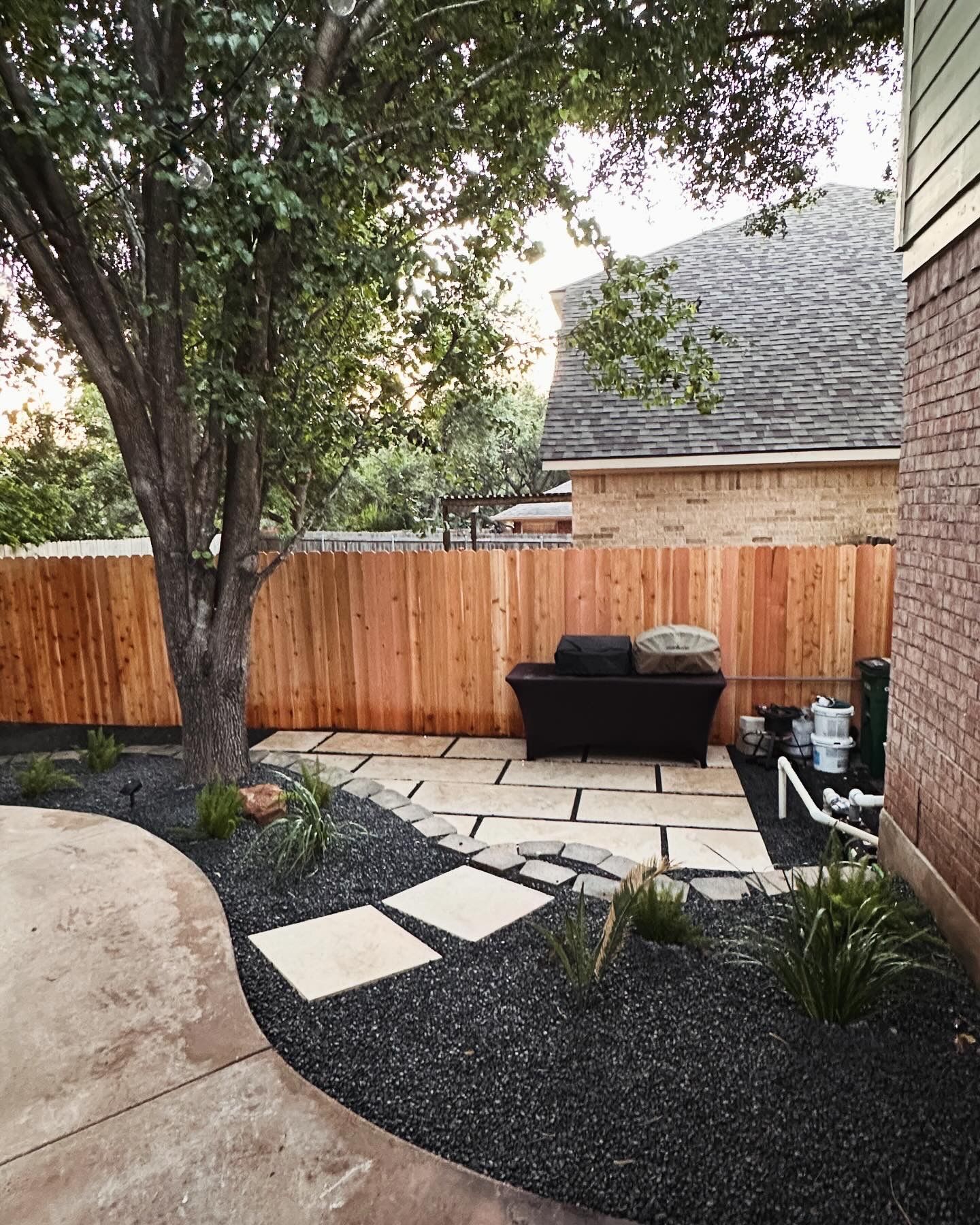 Backyard patio with stone path, black mulch, brown fence, and tree.