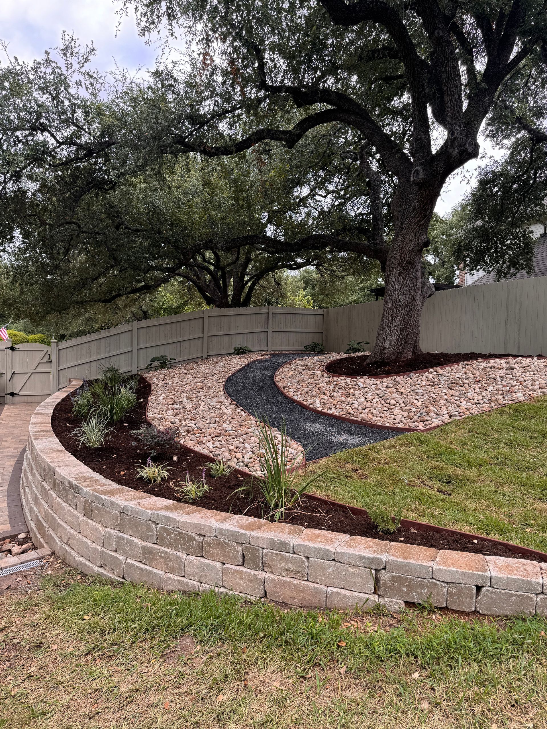 Curved brick retaining wall with a rock path leading to a tree, green grass, and a white fence.