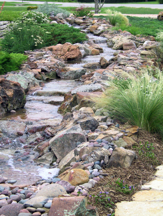 A stone-lined stream flows through a garden, with rocks and various plants along the banks.
