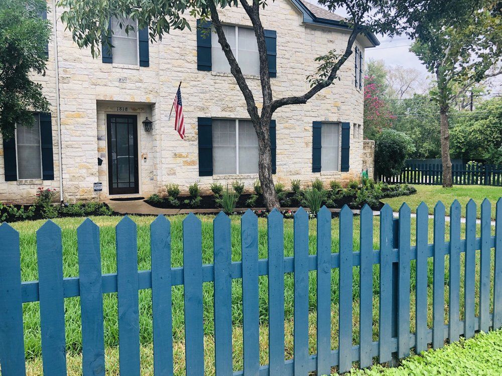 Blue picket fence in front of a two-story stone house with dark shutters and an American flag.