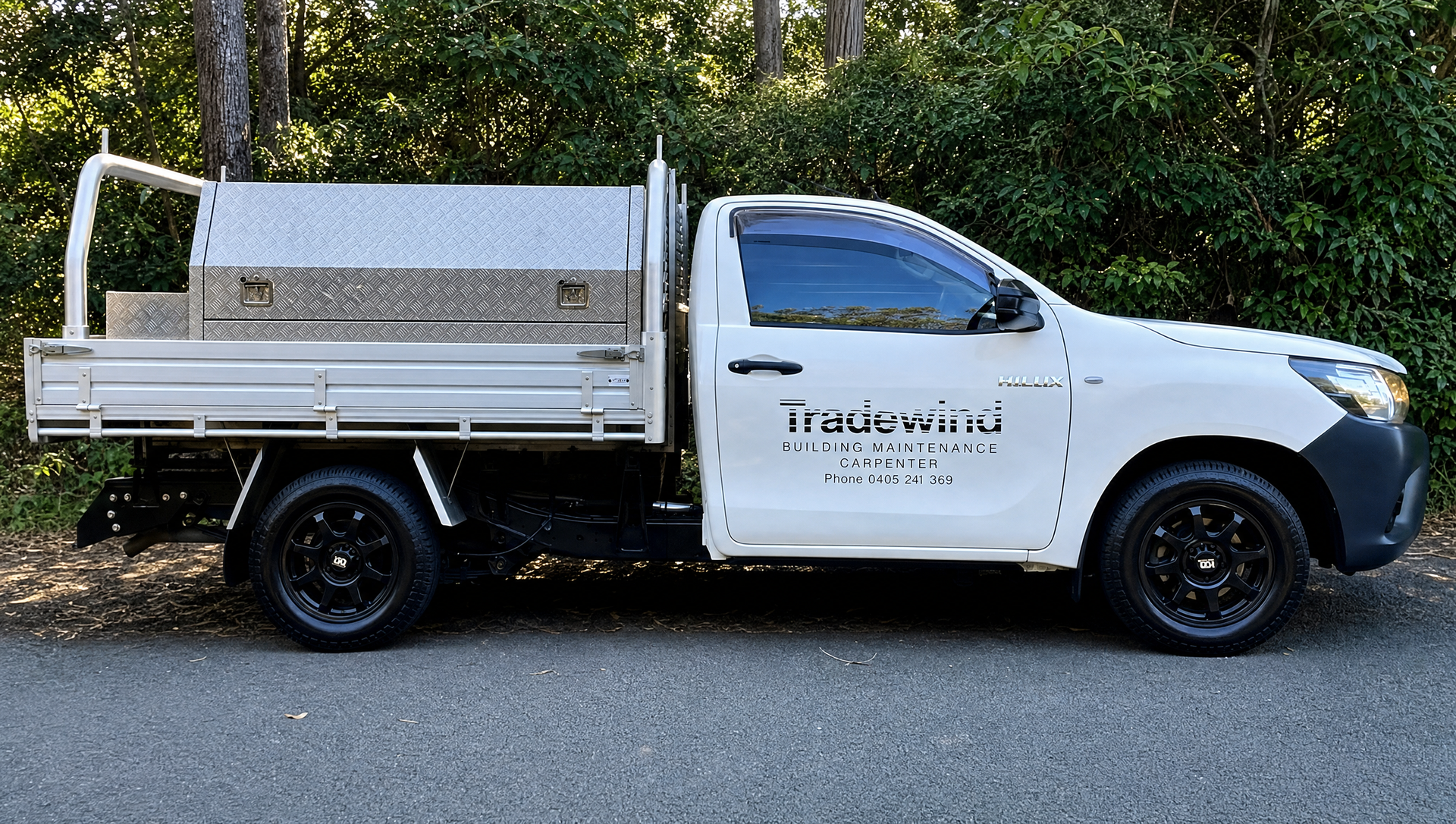 White utility truck with flatbed and sign on the door, parked on a roadside with trees in the background