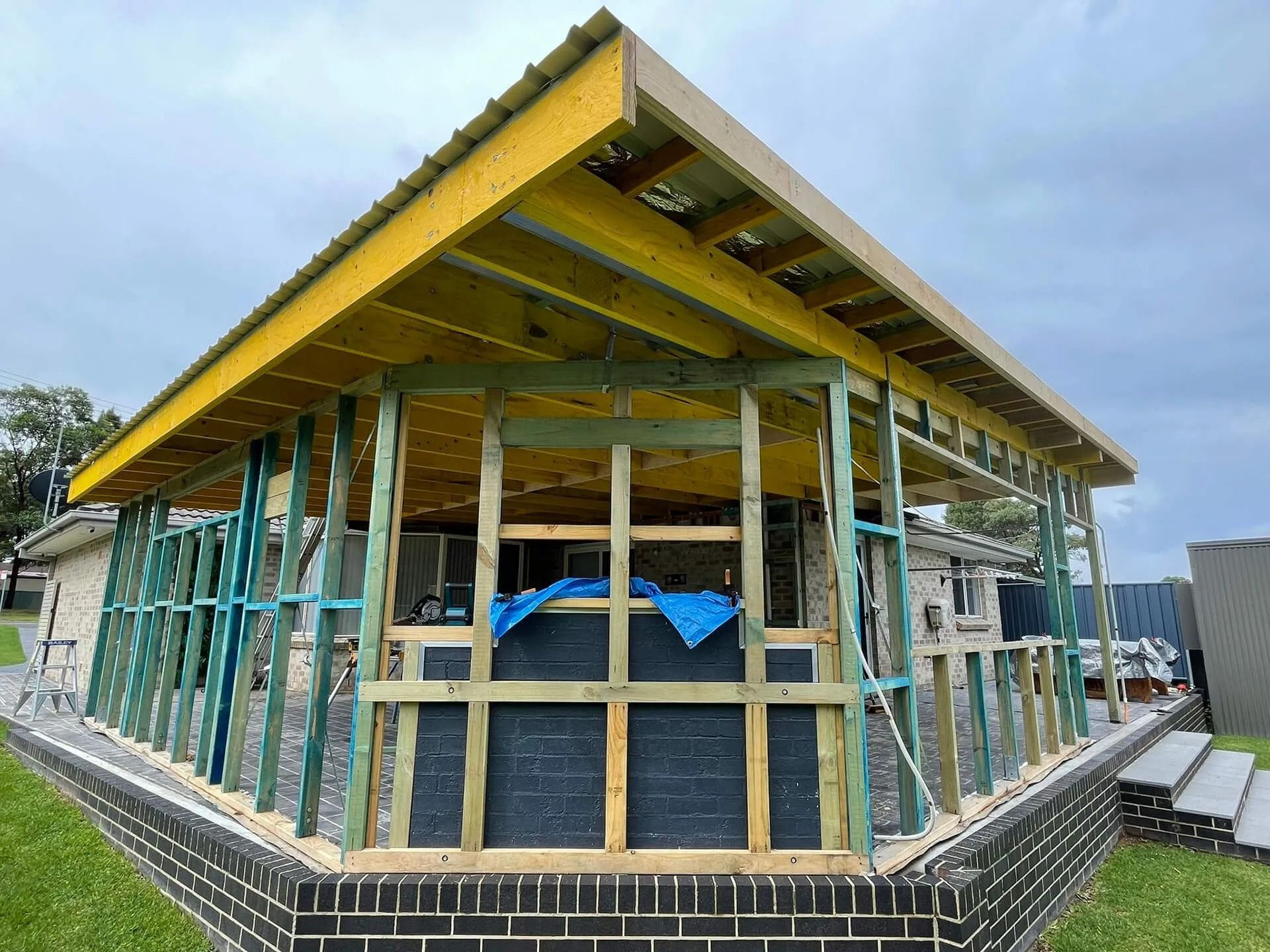 Partially built house with exposed wooden framing and green insulation wrap under a cloudy sky
