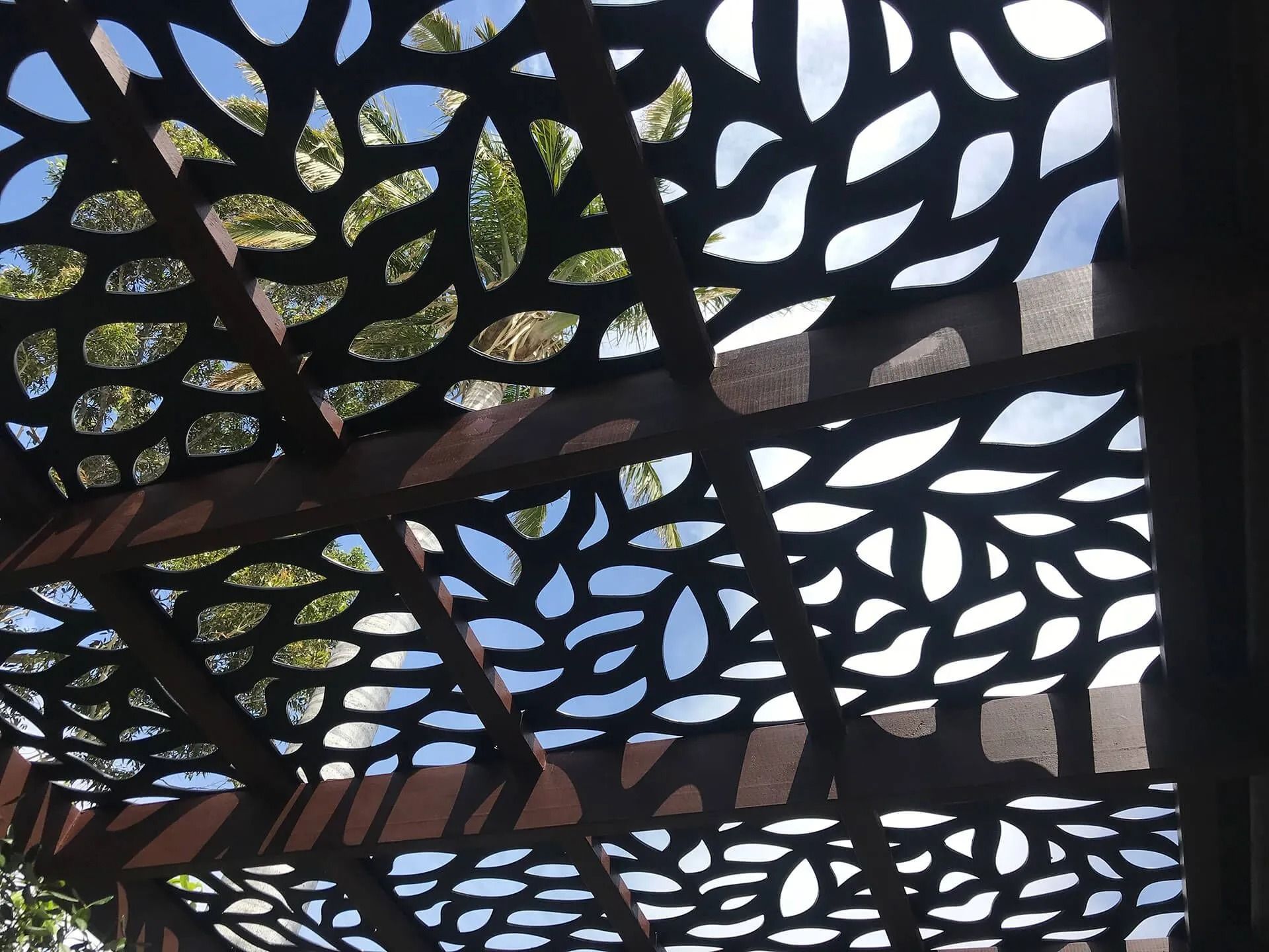 Black lattice overhead with organic cutout pattern against blue sky and trees
