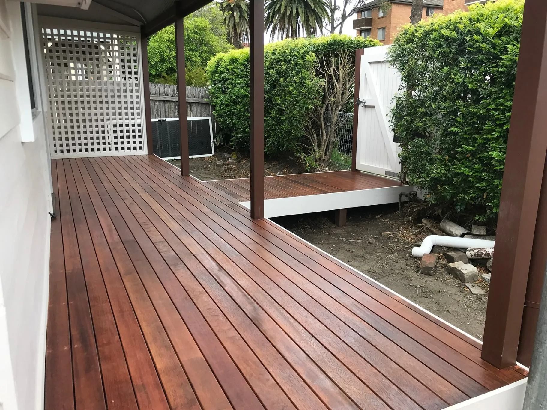 Covered wooden deck walkway beside a gravel yard, with white lattice and green hedges along the side.