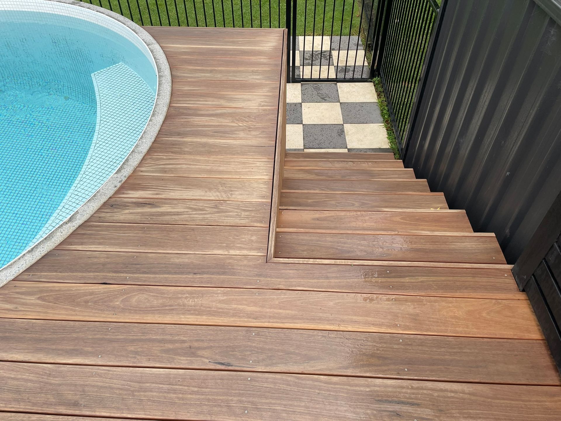 Wooden pool deck with steps down to a black gate and checkerboard patio tiles