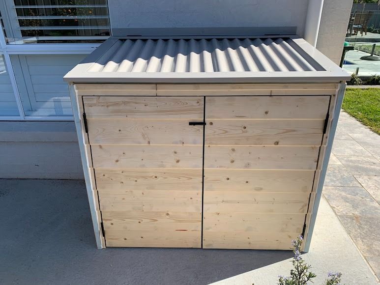 Wooden outdoor storage cabinet with double doors and a corrugated metal roof beside a building.