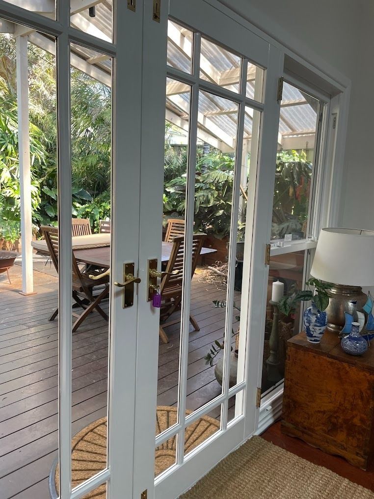 View through white French doors to a covered patio with wooden deck, garden chairs, and lush greenery