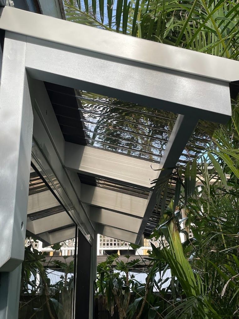 White pergola walkway seen from below, framed by tropical plants and sunlight.