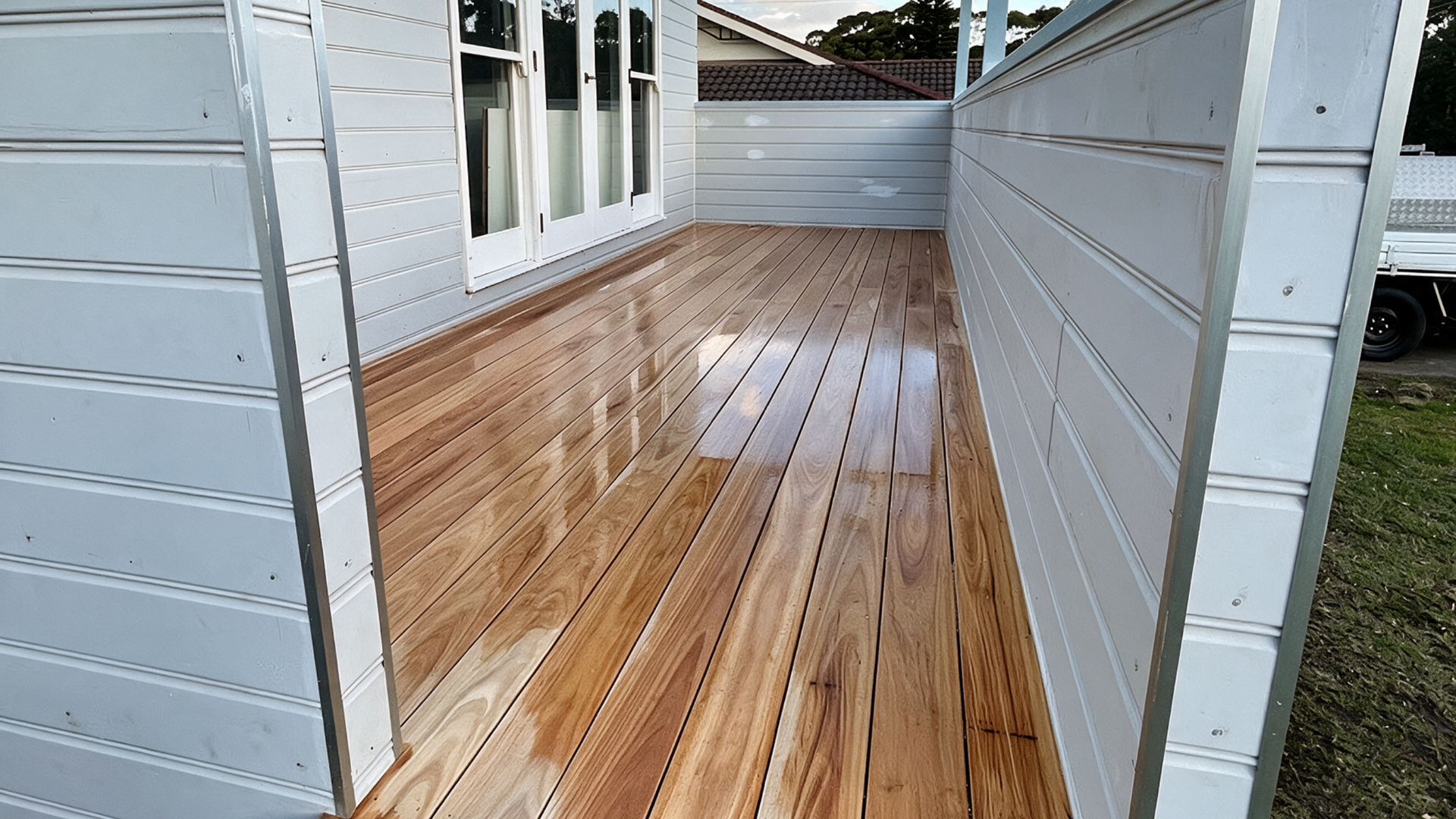 Narrow wooden deck walkway between two white buildings, with windows and a shiny floorboards finish.