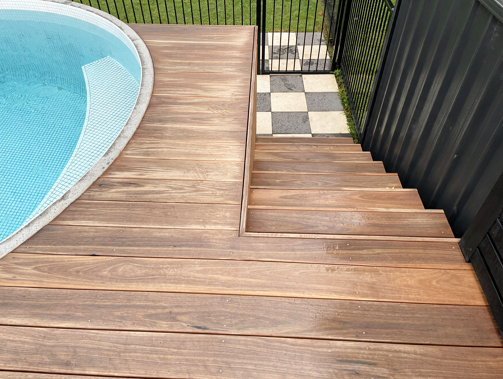 Wooden deck stairs beside a pool, leading to a black gate and checkerboard patio tiles.