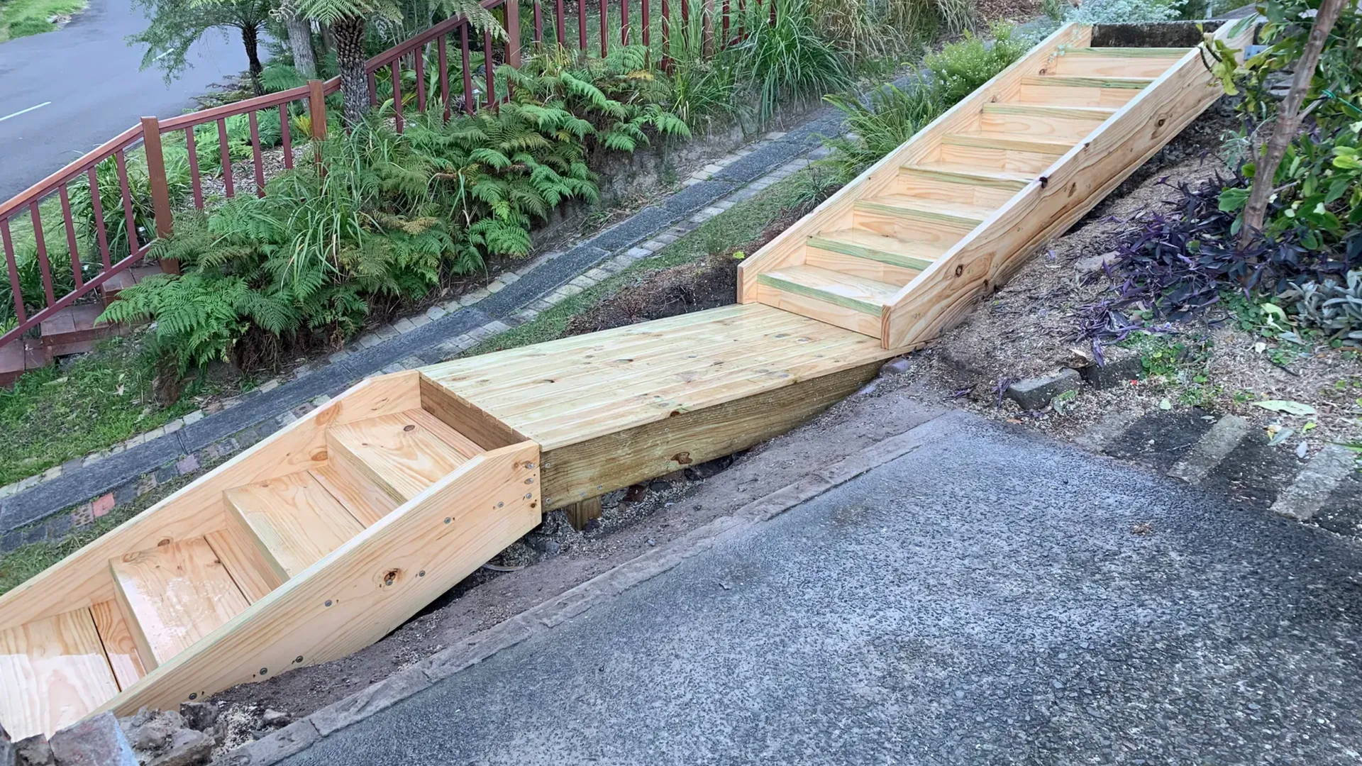 Wooden ADA-style ramp and steps built along a sloped roadside beside a fence and greenery