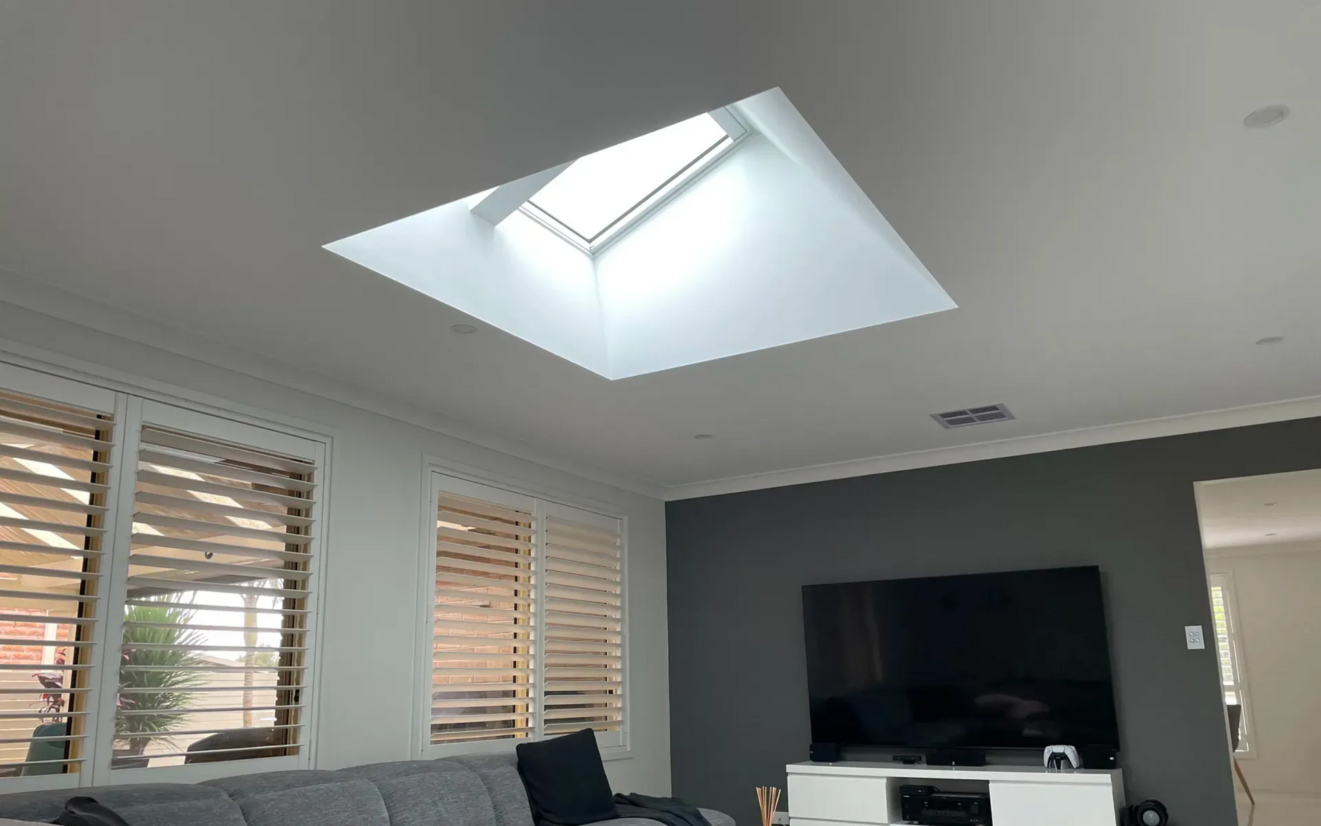 Bright living room with skylight, white blinds, gray accent wall, and a large TV on a stand.