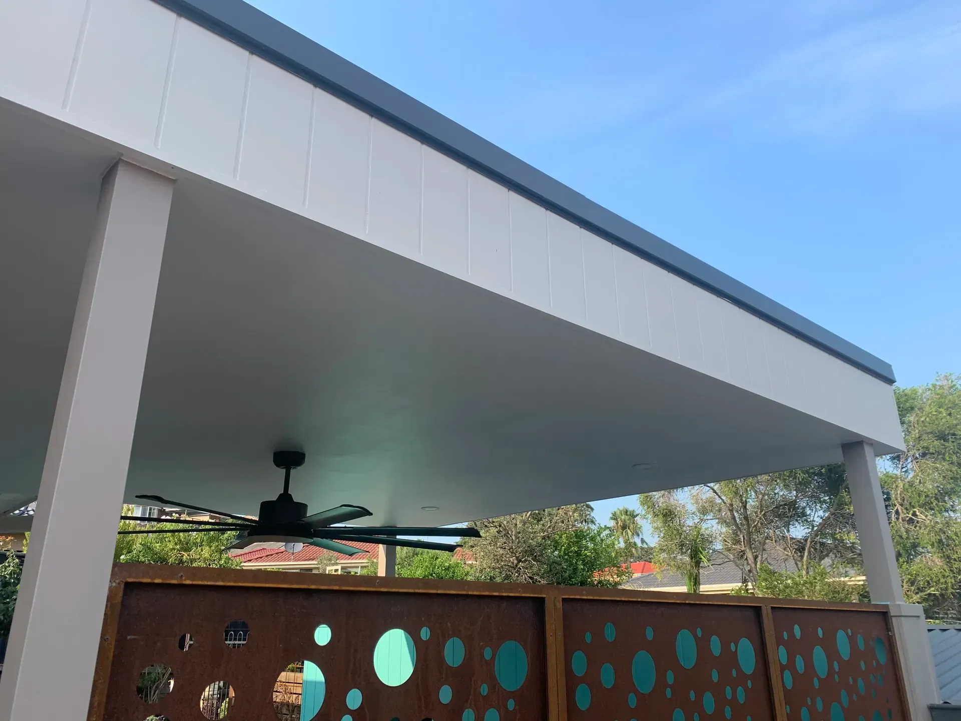 Modern patio with white roof, ceiling fan, and brown privacy fence with circular cutouts under a blue sky