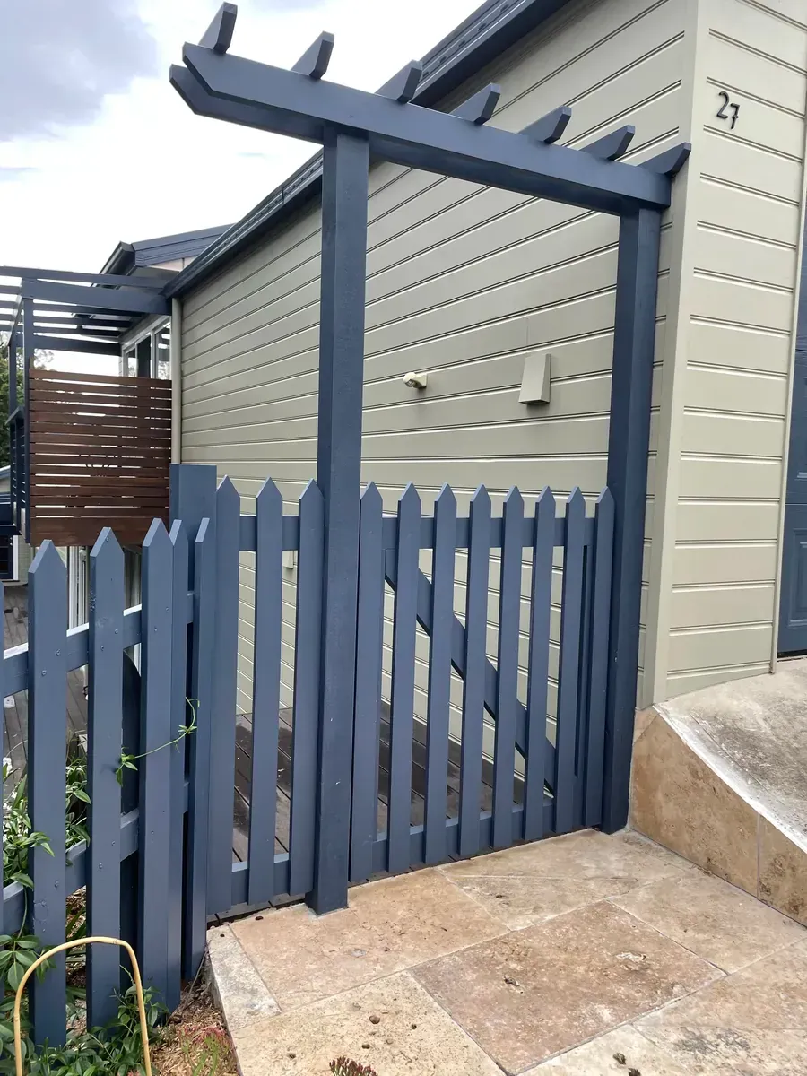 Blue wooden picket gate and pergola beside a beige house with a concrete path