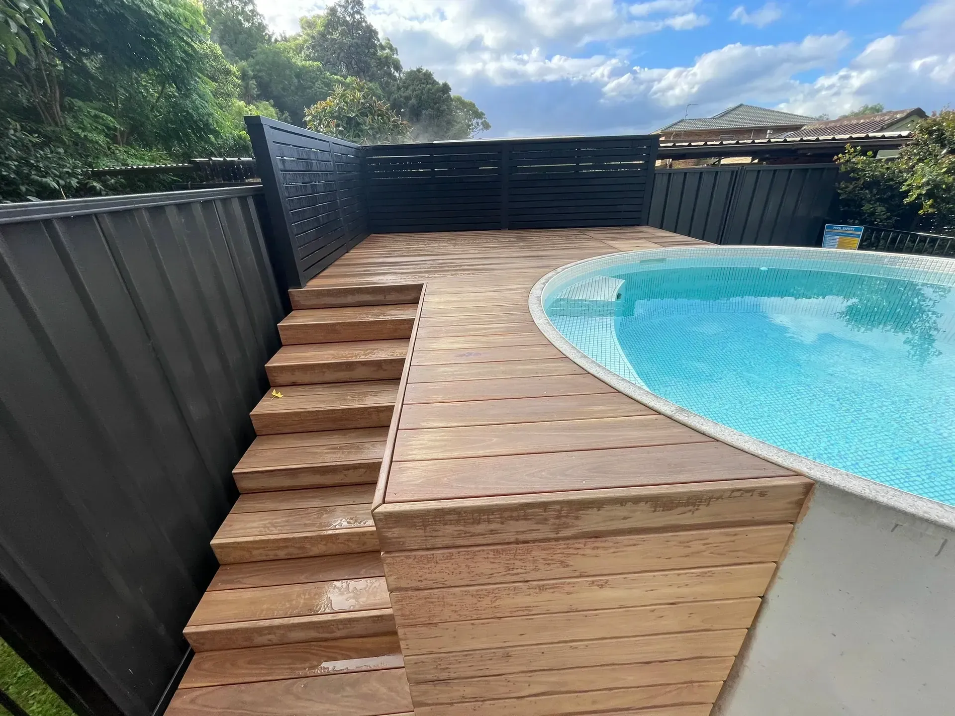 Wooden pool deck with steps beside a curved blue swimming pool and black fence.