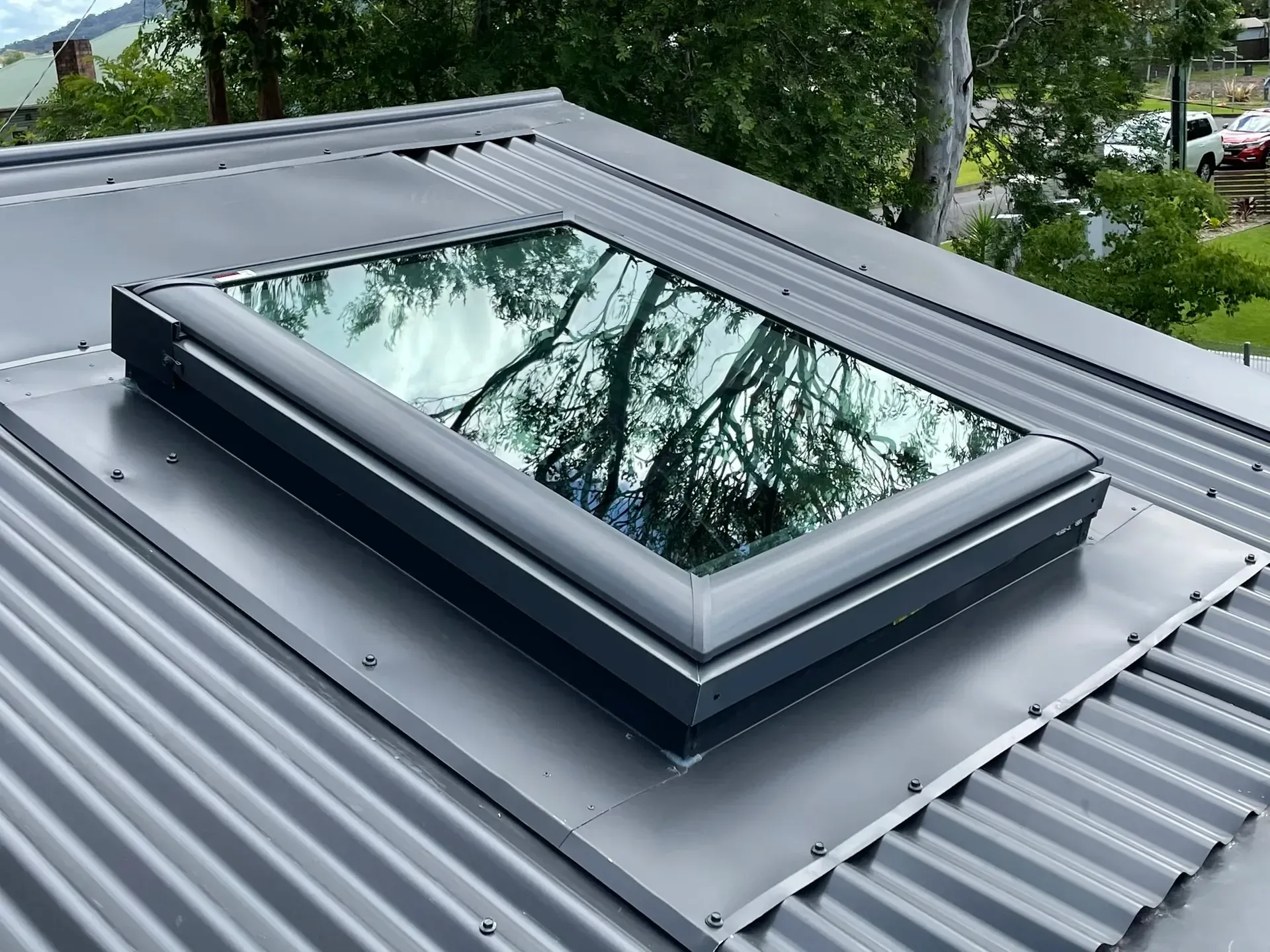 Rooftop skylight with dark frame set into a gray metal roof, reflecting trees and sky