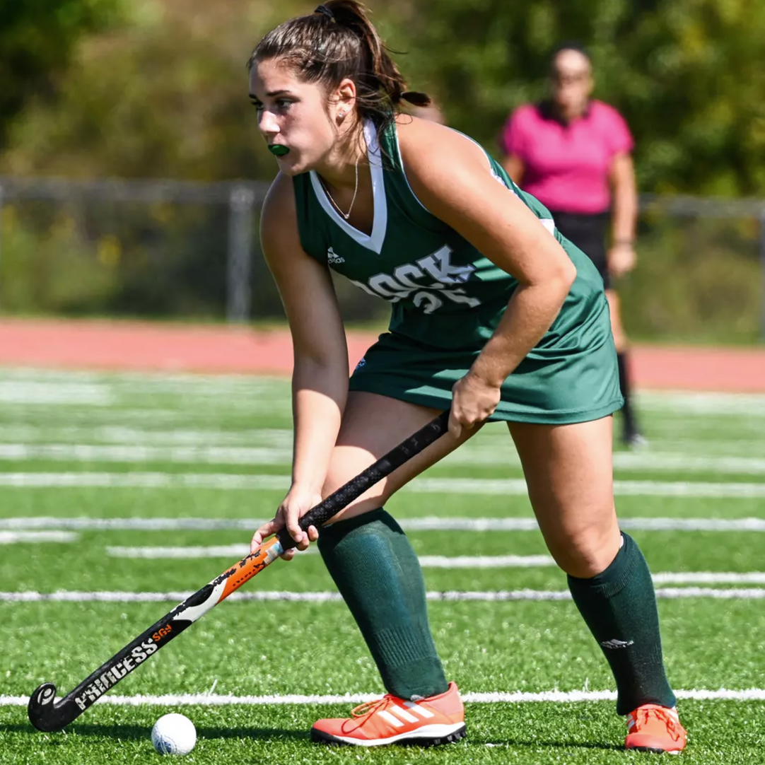 A player in a green uniform focuses on a field hockey ball while holding a stick on a turf field.