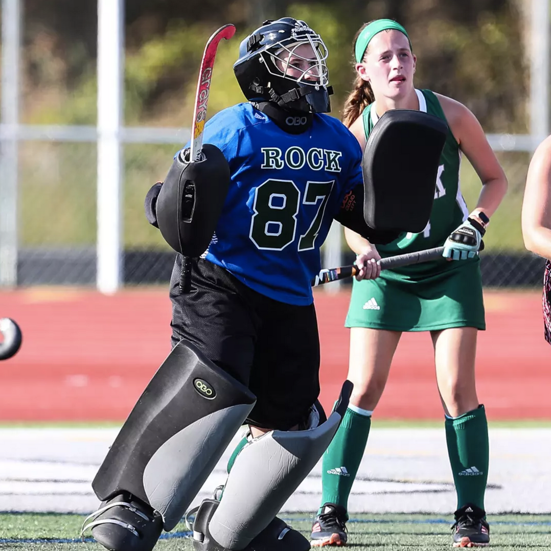 A field hockey goalie in a blue jersey and protective gear stands on a field near a player in a green uniform.