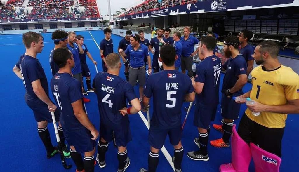 Men's field hockey team in a huddle on a blue field, wearing navy blue jerseys, with the coach in the center.