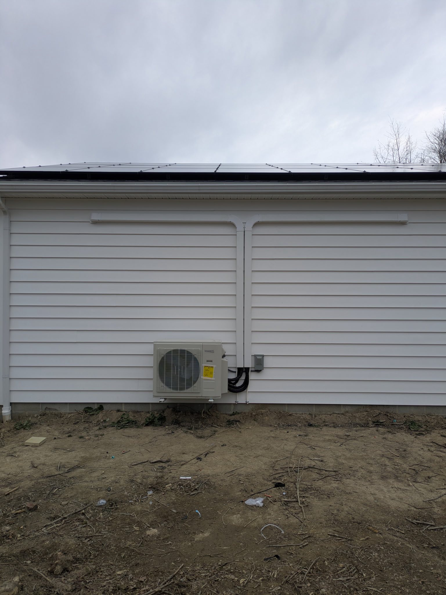 White-sided garage with closed doors, outdoor AC unit, and bare dirt foreground under cloudy sky