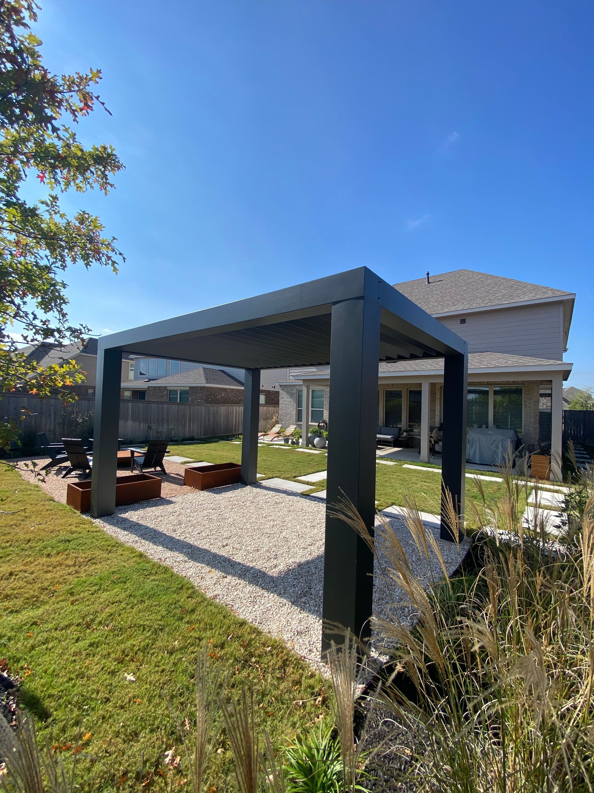 Black pergola in a backyard with gravel, grass, and a house in the background under a blue sky.
