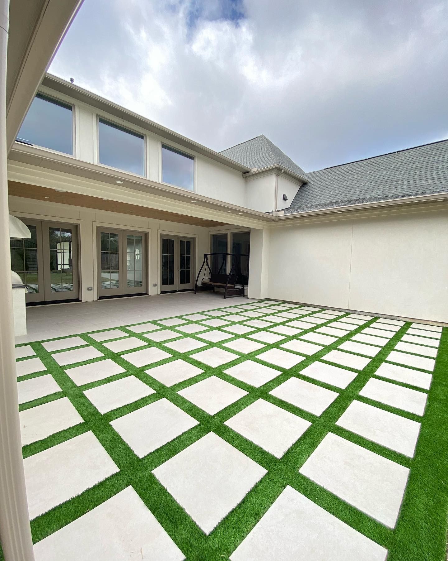 Backyard patio with square white pavers interspersed with green turf, surrounded by a white house and a cloudy sky.