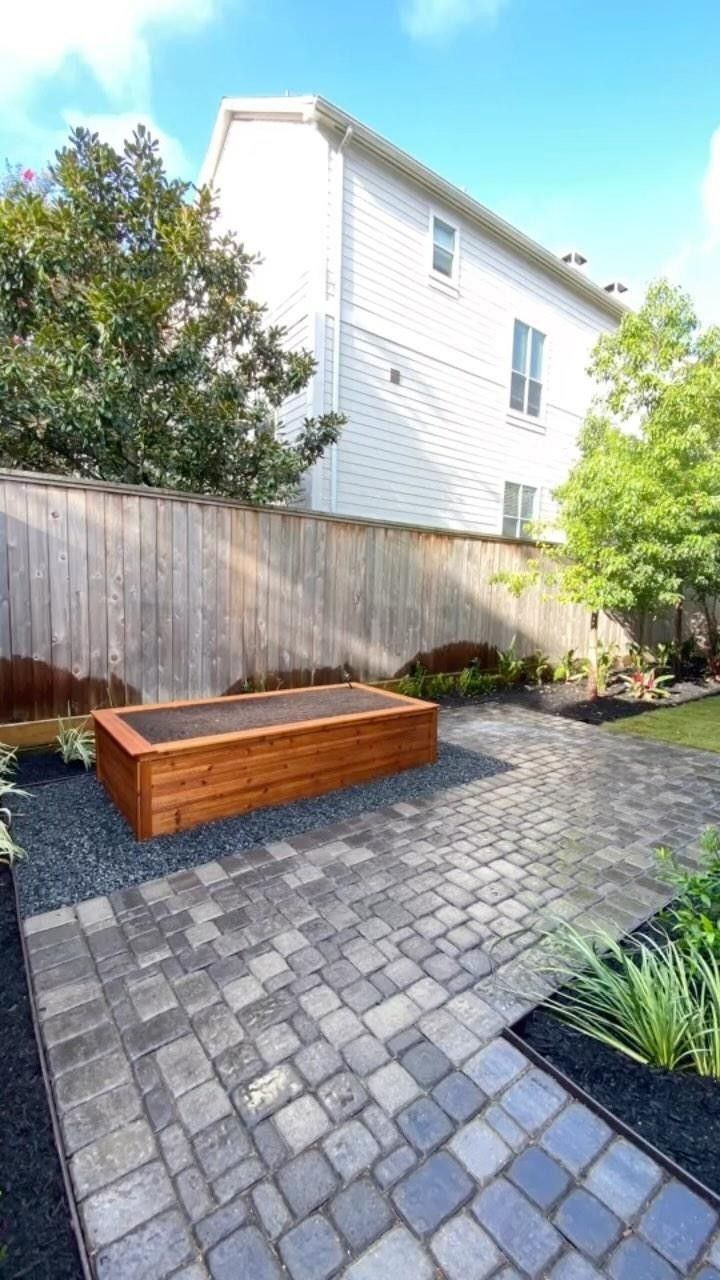 Backyard with a wooden planter box on a brick patio, a wooden fence, and a two-story white house in the background.
