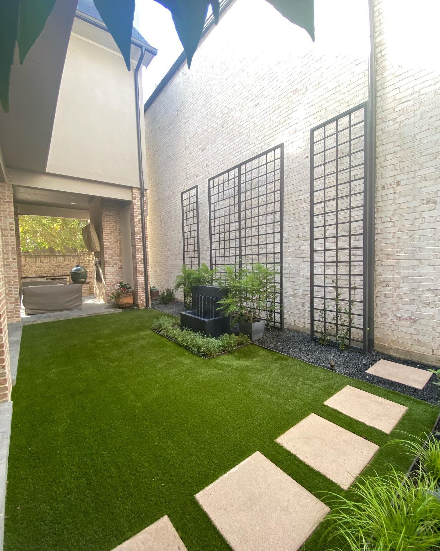 A modern backyard with artificial turf, stone path, trellis against a brick wall, and a fountain.