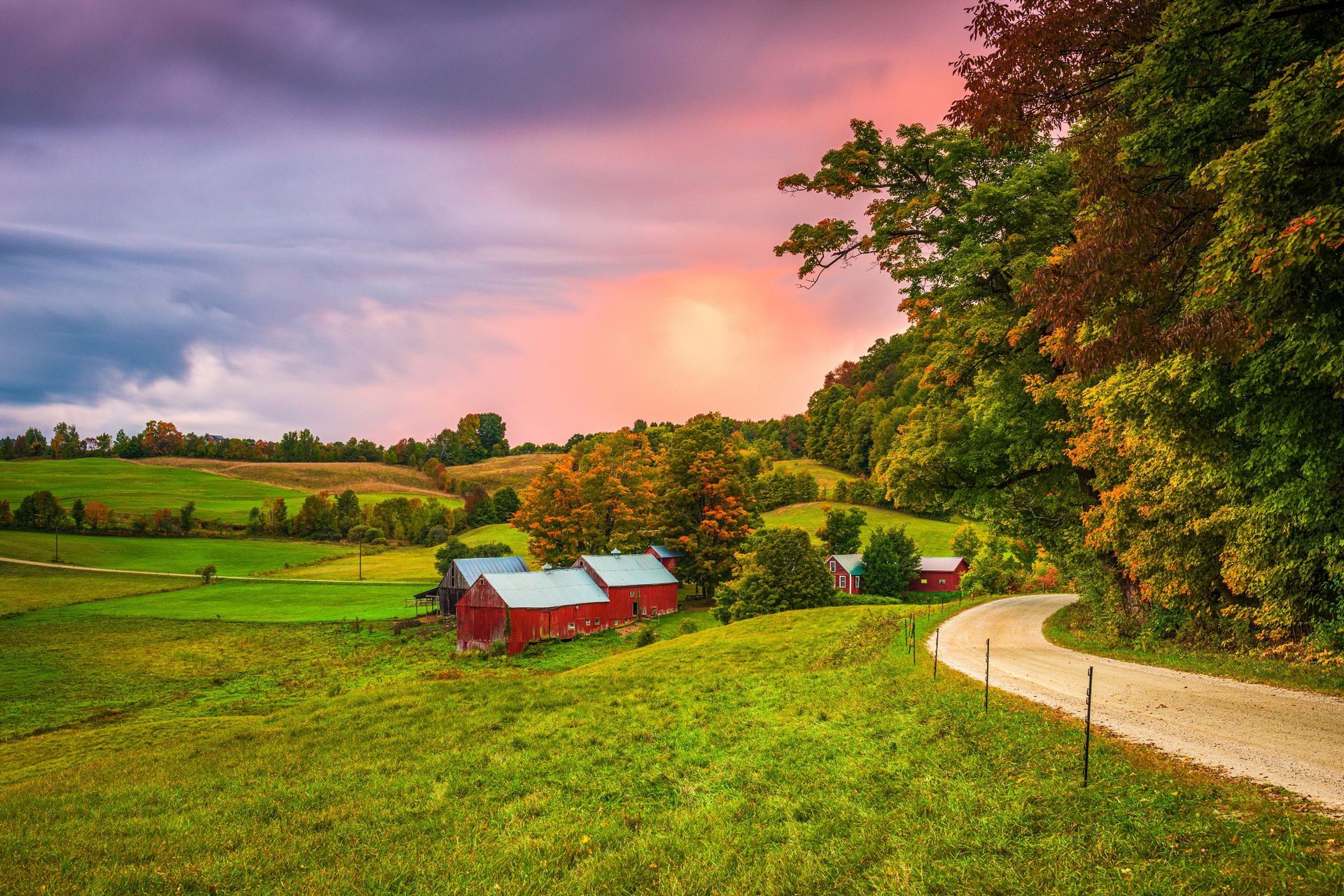 Red barns sit in a rolling green field beside a dirt road under a dramatic, colorful sunset sky.