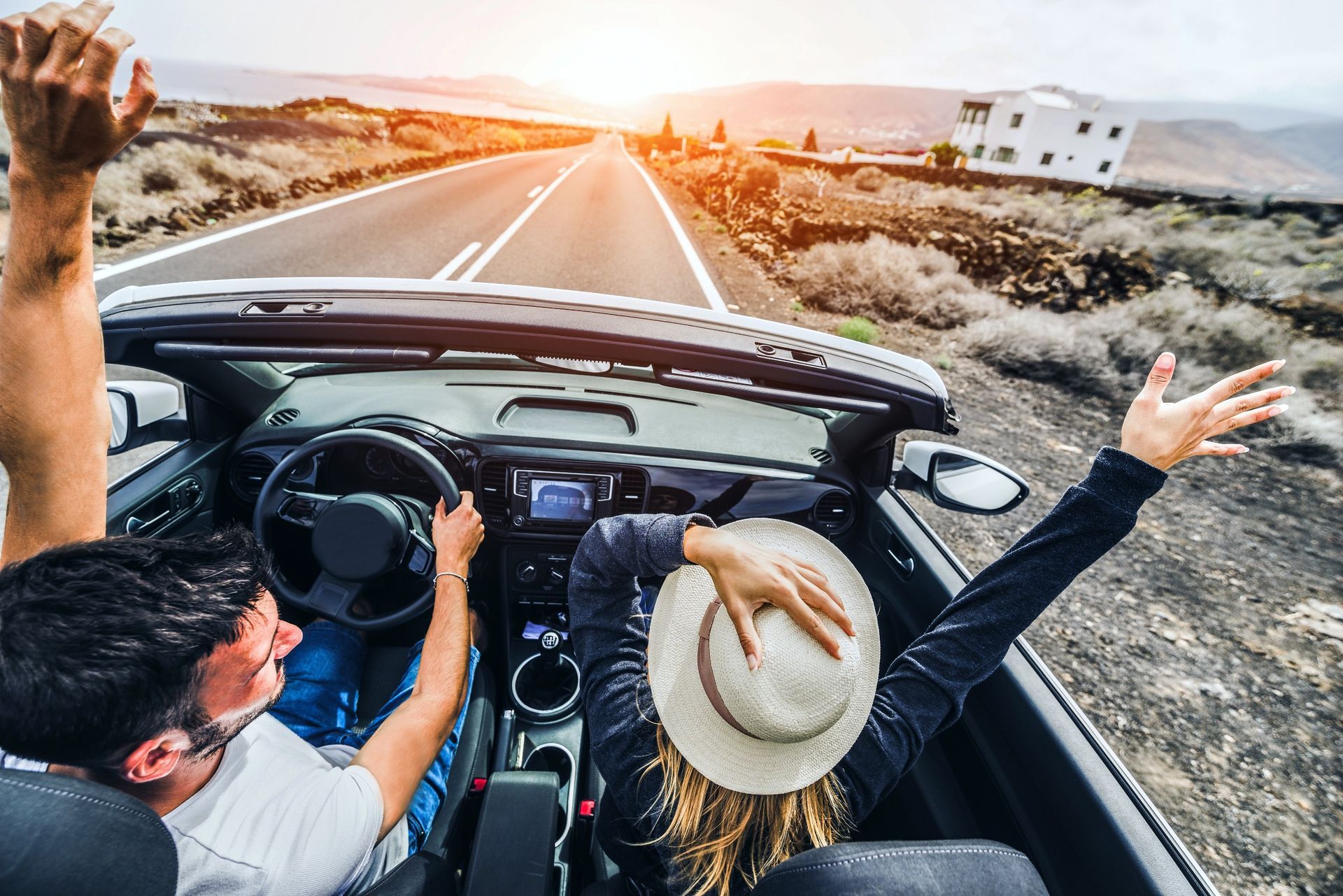 Two people in a convertible car drive down a scenic road with their arms raised in celebration.