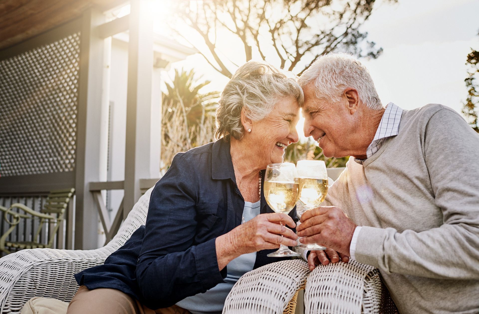 Two people sitting close together on a wicker sofa, smiling and clinking wine glasses in a sunlit outdoor setting.