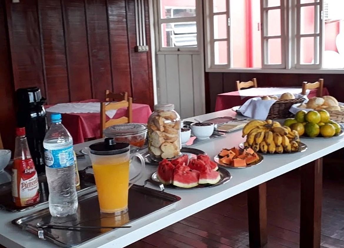 Mesa com frutas, bebidas e petiscos disposta em uma sala de madeira junto a uma janela.