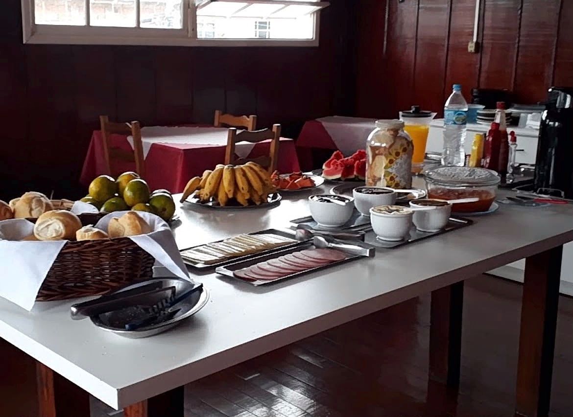 Mesa de buffet com frutas, pães, bebidas e conjunto de chá em uma sala de jantar.