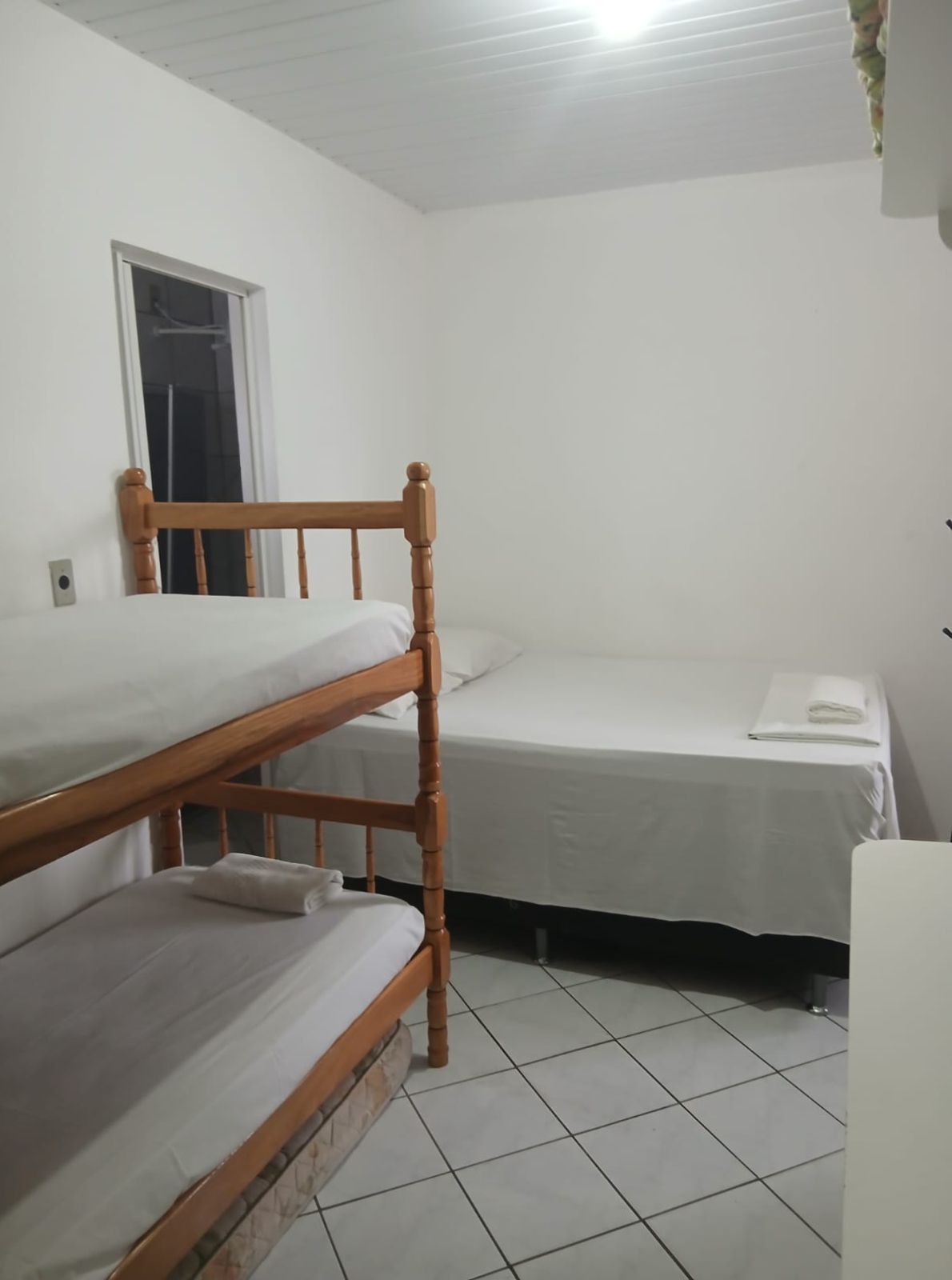 Quarto pequeno com um beliche de madeira, uma cama de solteiro, piso de azulejo e uma janela.