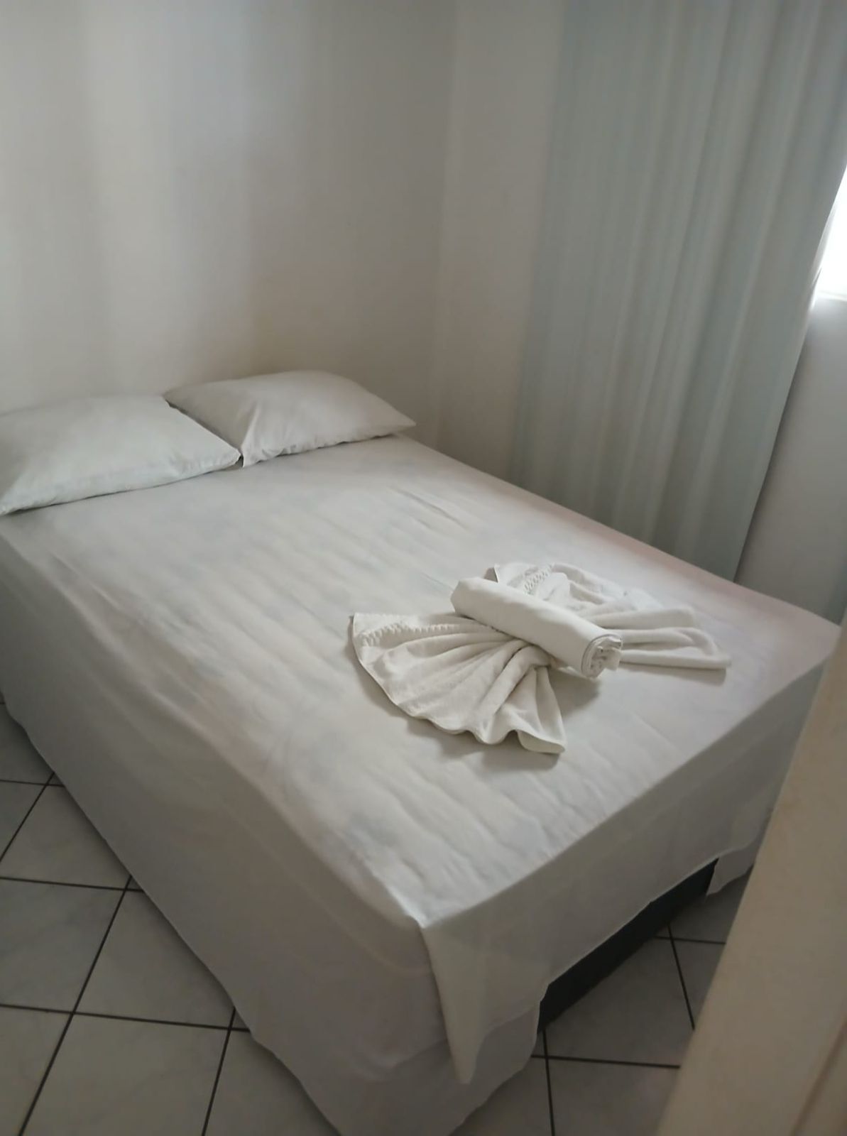 Cama branca arrumada com toalhas dobradas em um quarto claro com azulejos, ao lado de cortinas translúcidas.