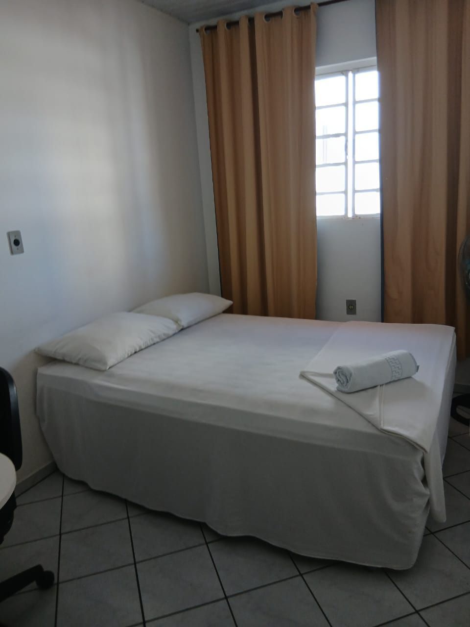 Quarto pequeno com uma cama de solteiro, lençóis brancos, cortinas bege e uma janela ao lado da cama.