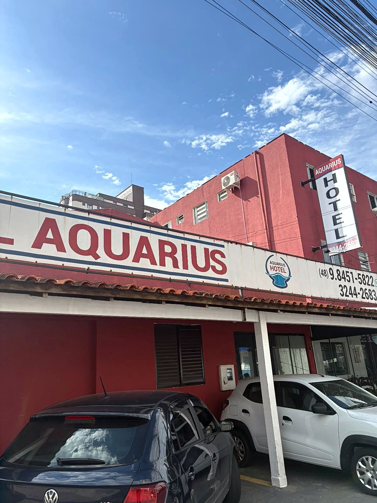 Fachada vermelha do hotel Aquarius com carros estacionados sob um céu azul.