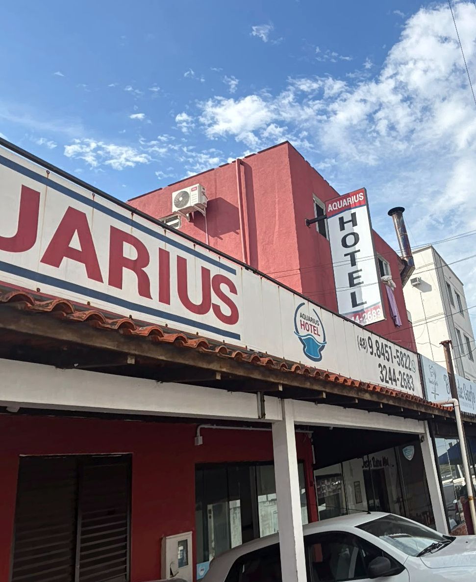 Vista da rua do Hotel Jarius com fachada vermelha e branca sob um céu azul, com carros estacionados em frente.