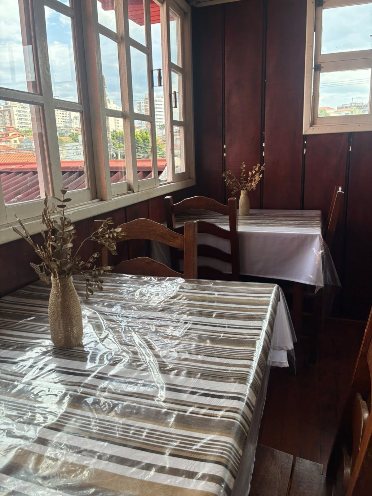Pequena sala de jantar com toalhas de mesa xadrez, cadeiras de madeira e janelas com vista para os telhados.