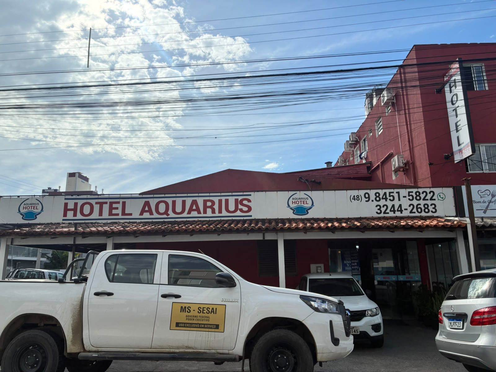Fachada do Hotel Aquarius com veículos estacionados sob um céu nublado.