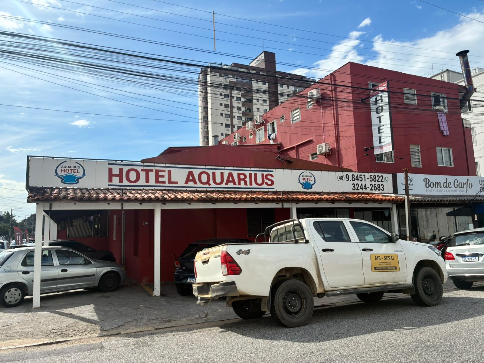 Fachada do Hotel Aquarius com prédio vermelho, SUV branco estacionado em frente e fios elétricos suspensos.