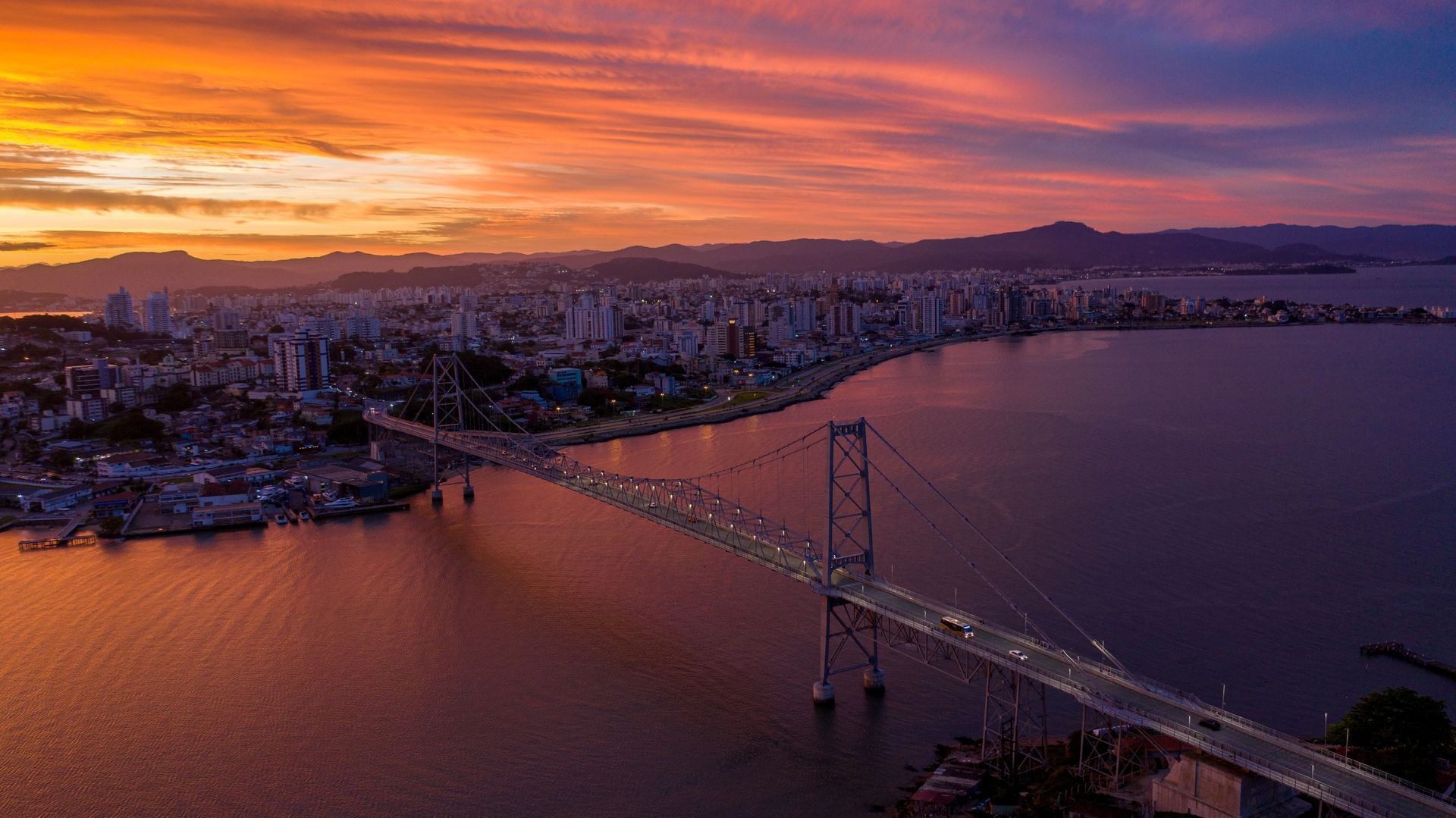 Pôr do sol sobre uma cidade costeira com uma ponte suspensa iluminada atravessando a baía.