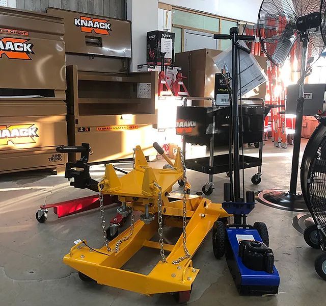 Yellow snow plow attachment and blue floor jack in a garage with shelves and tools.