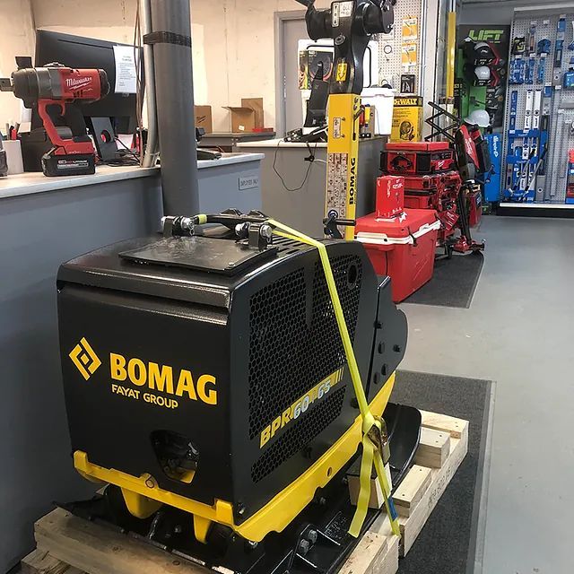 Black BOMAG construction machine strapped on a pallet in a workshop, with tools and equipment in the background