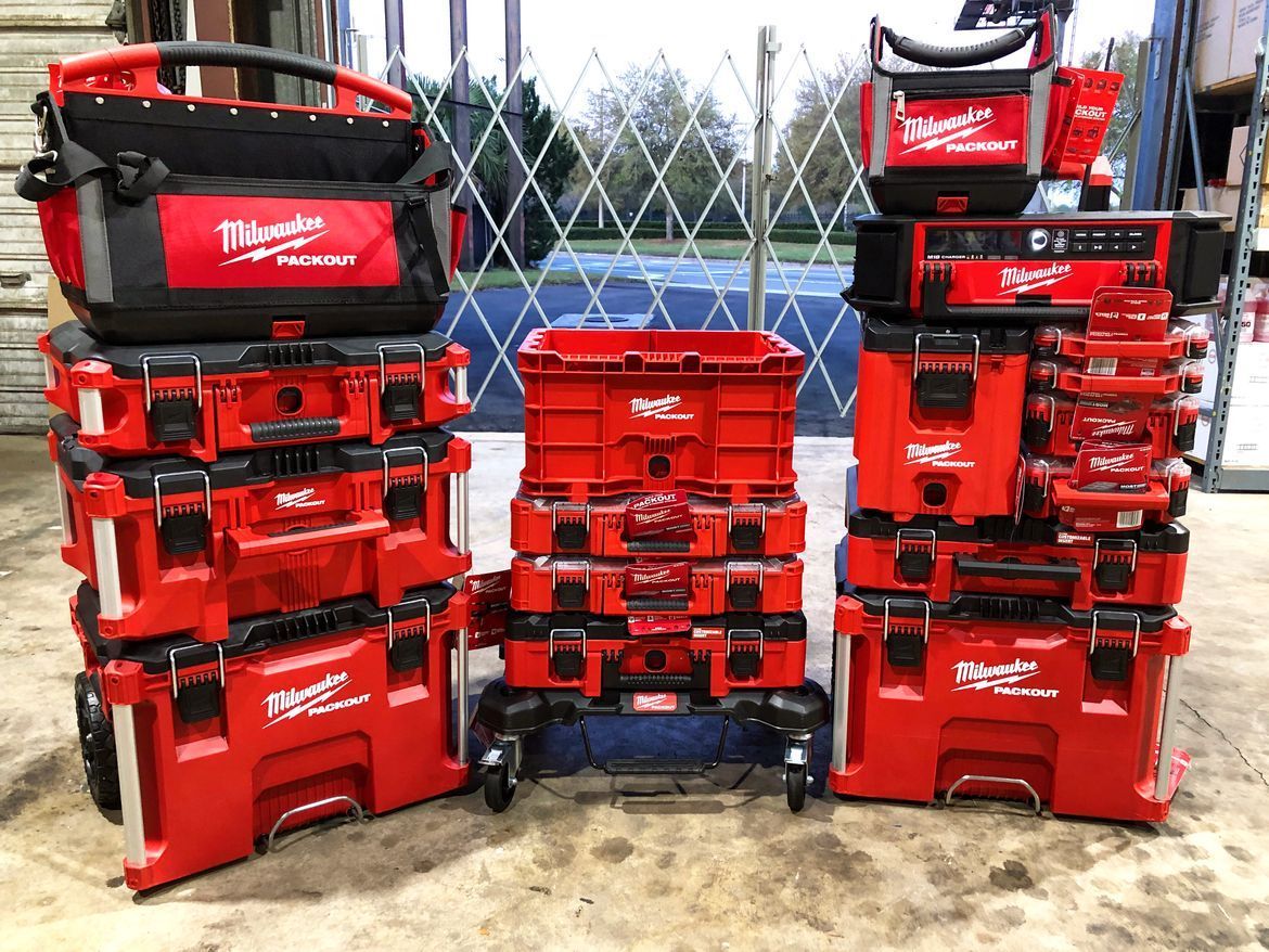 Stacks of red Milwaukee tool bags and storage boxes displayed outdoors in front of a fence