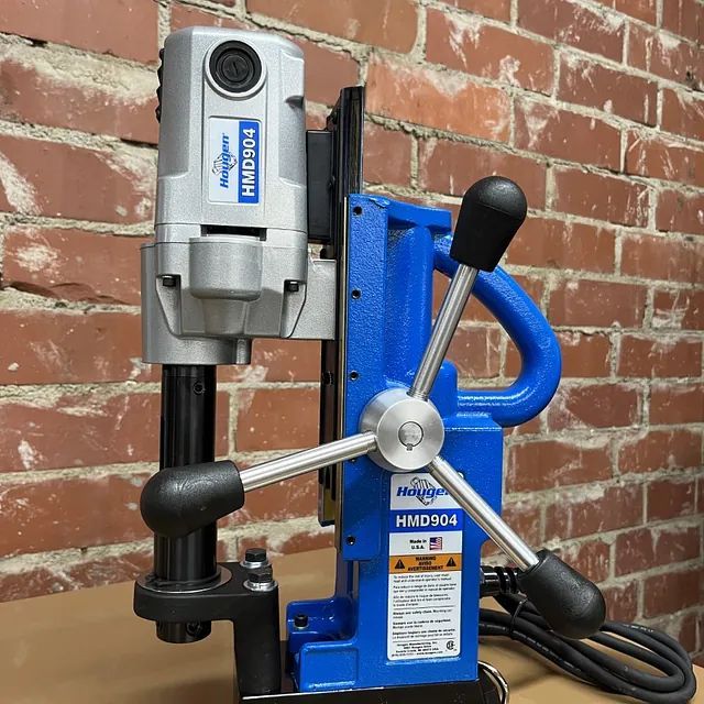 Blue magnetic drill press with silver motor mounted on a black base against a brick wall